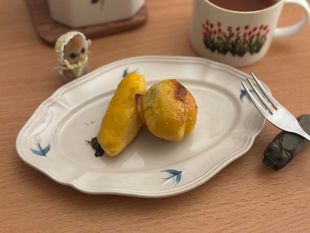 ツバメの模様の入ったオーバルの皿にスイートポテトが2個のっている。後ろにはティーポットとカップ。右側に木彫りの熊の箸置きに立てかけたフォークが置いてある。
シルバニアのミーアキャットの赤ちゃんがスイートポテトに近づいている。
