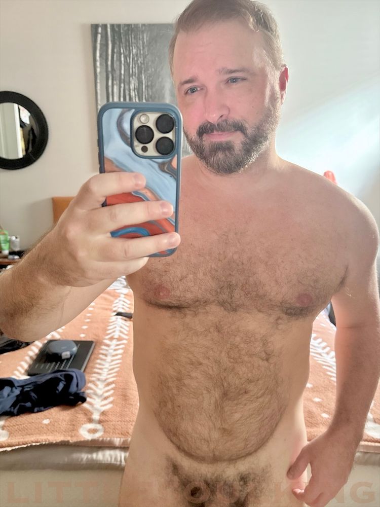A selfie of a furry, bearded man standing naked in front of a mirror in his bedroom with his bed directly behind. 