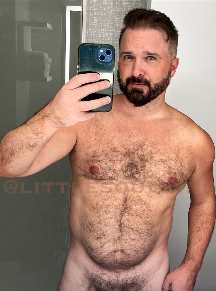 A hotel mirror selfie of a completely naked, furry, bearded man. 
