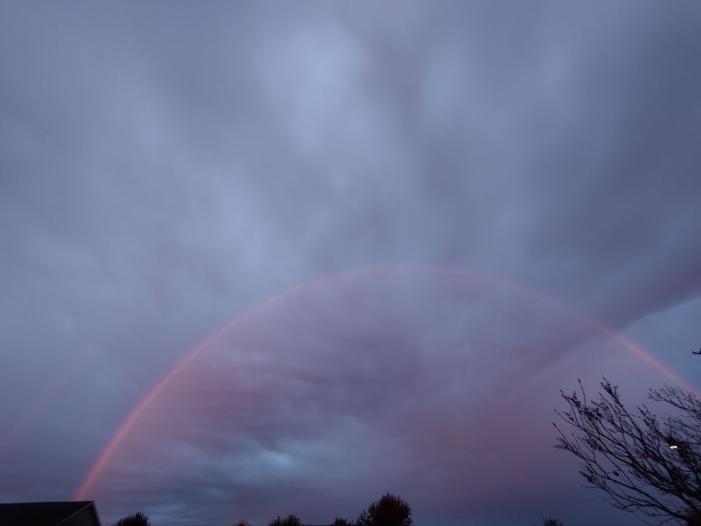 The photograph shows a primary rainbow with a faint secondary rainbow visible. The setting is a very overcast morning with low-hanging cumulus clouds with the peaks of houses and trees barely visible in the bottom of the frame.
