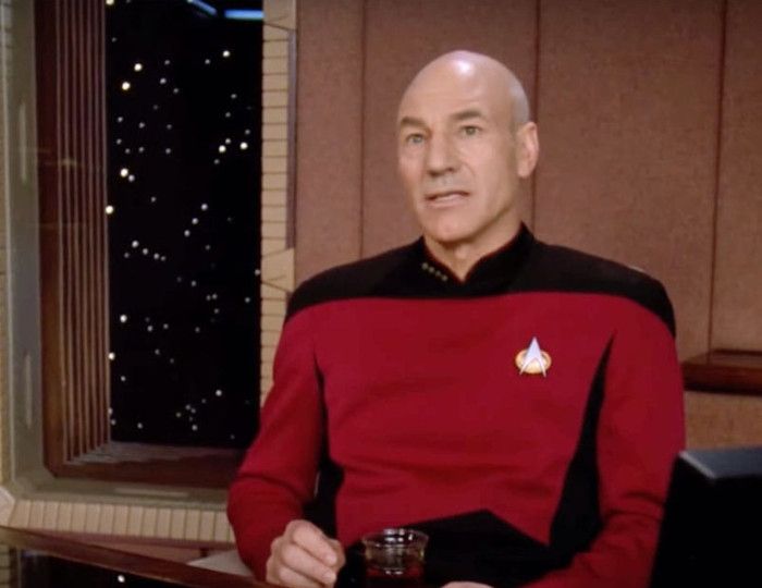Captain Picard sits at his desk in the ready room with the window looking out at the stars behind them, wearing a thoughtful look as he holds a cup of tea.
