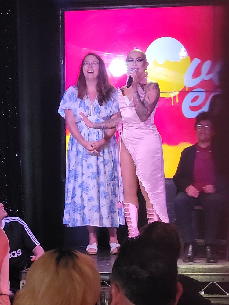 Ongina in a pink dress on stage with a young woman in glasses, long brown hair, and a long floral print dress.