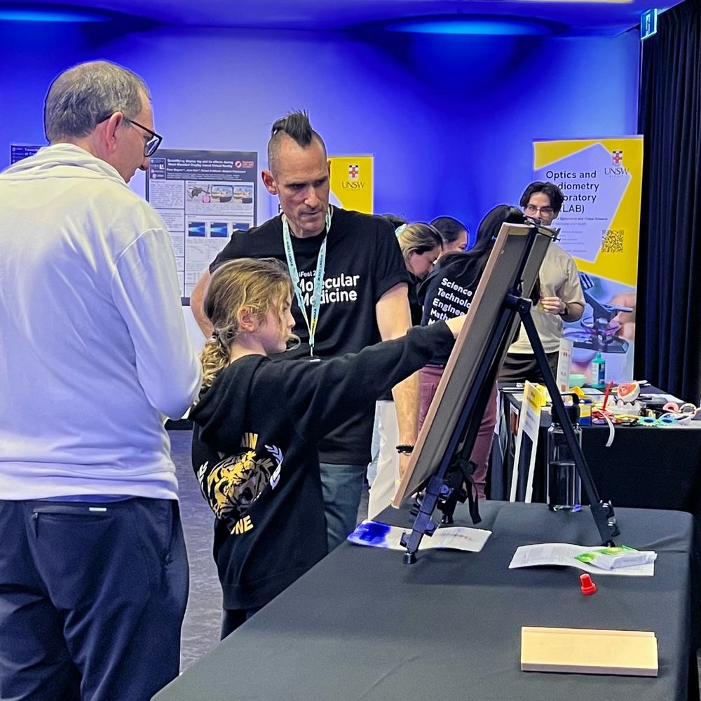 A father and daughter speaking with Dr Jon and pinning the superhero capes on the art board game of bacteria deployed to save the Great Barrier Reef.