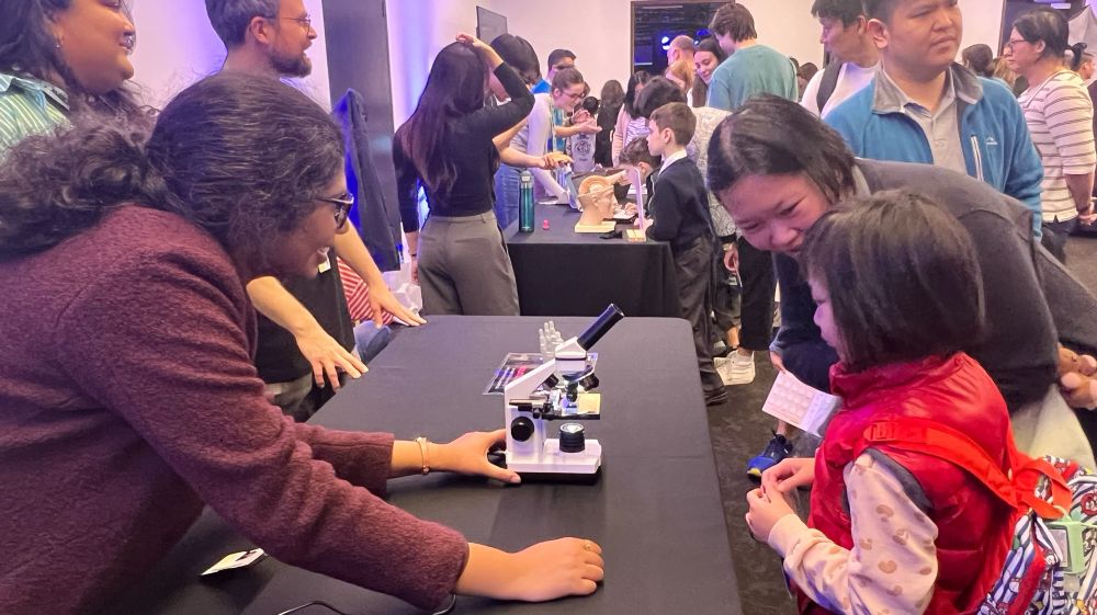 A conversation between Upasana from Molecular Medicine and a little girl and her mum. Upasana is leaning over a table with a miniature microscope.
