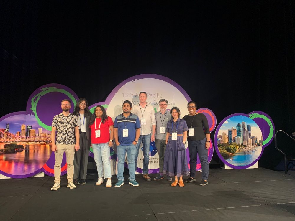 Group photo of eight colleagues representing the dept, standing in front of the conference mural