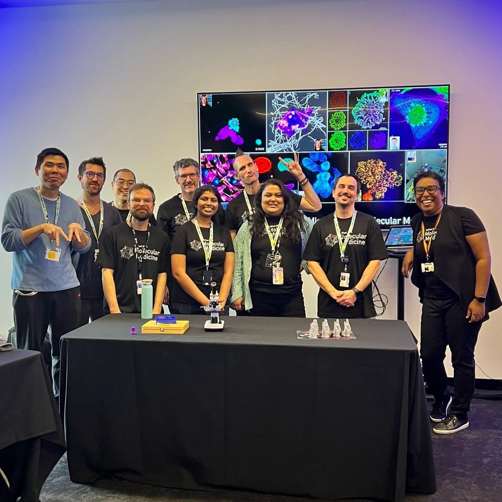 A group photo of the Molecular Medicine (Single Molecule Sciences) team. In front, a table with some objects, including a miniature microscope. In the background, a large tv screen with vibrant images of cells and molecules.