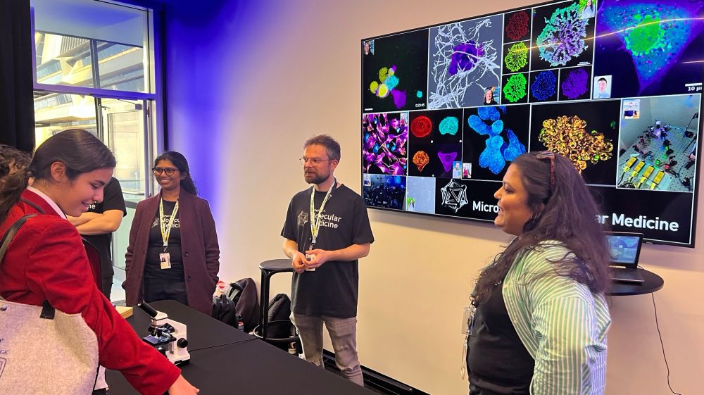 Some team members of Molecular Medicine standing in front of a colourful big tv screen and talking to some of the SciFest visitors