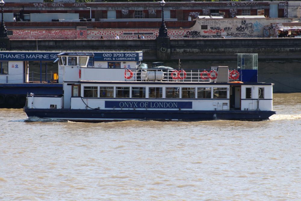 Popular party boat ONYX OF LONDON sails downstream on a greyish River Thames past the Thames Marine Services' fixed refuelling barge and the National Covid Memorial Wall. The yellow deck chair is just visible....