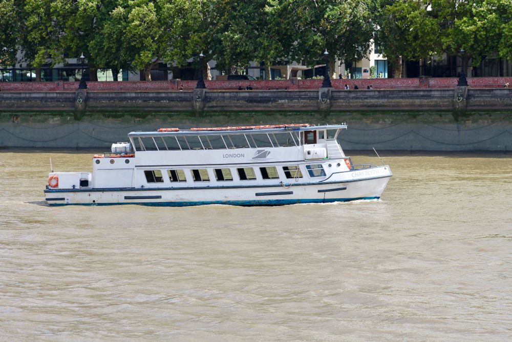 Newly re-painted, white Thames party boat CHAY BLYTH heads upstream along Lambeth Reach, past the thousands of red hearts of the UK National Covid Memorial Wall and the green overhanging branches of London plane trees.