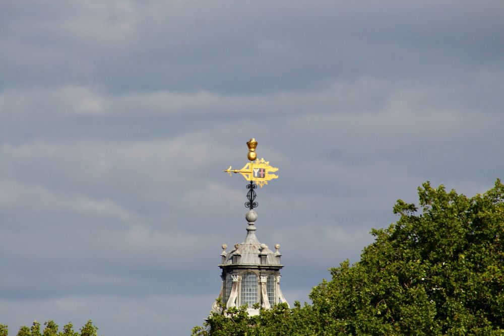 Bright golden weather vane pointing north, resting on a glass and light grey stone lantern, with tops of plane trees on the bottom right of the picture. If you would like to read my article on Lambeth Palace, see: https://thetidalthames.com/2023/06/25/lambeth-palace-from-the-river/ 