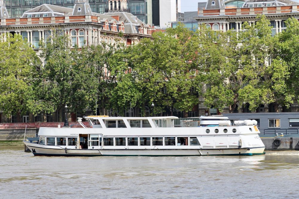An elegant white boat leaving her tree-lined mooring. Beyond are the *Italianate* buildings of part of St. Thomas' Hospital in London.