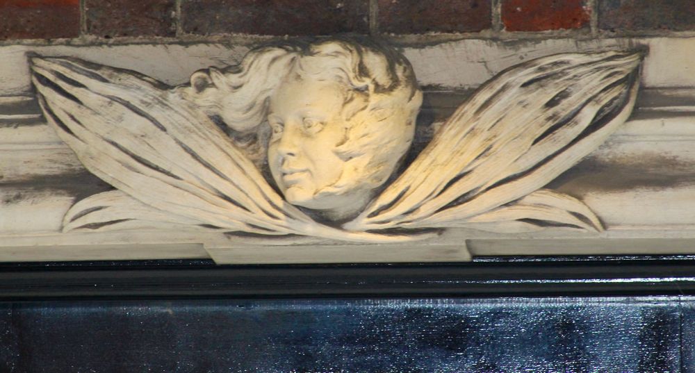 A sculpted angel head and wings, caught in a light, watching over a doorway.
