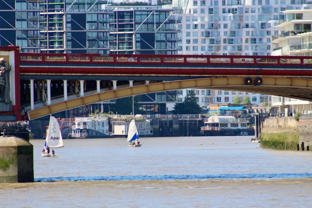 Two white sailing boats tacking across the river Thames beyond the yellow and reddish brown colours of Vauxhall Bridge.
