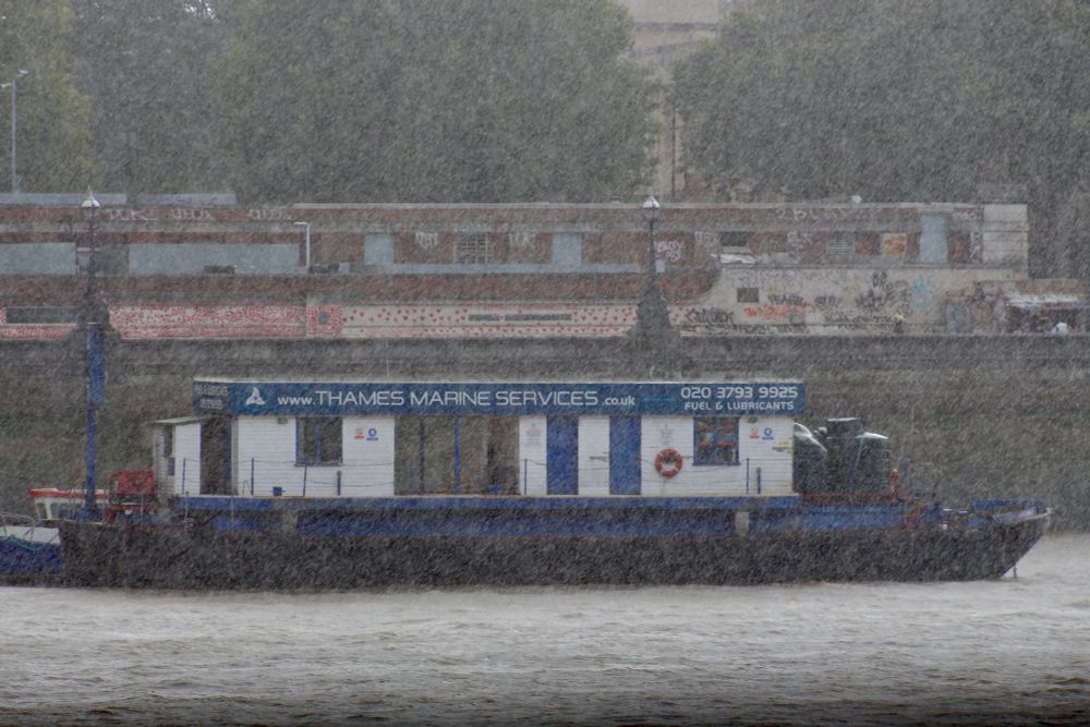 Rain-blurred view of blue and white Thames Marine Services' refuelling barge moored below the red hearts on the UK's National Covid Memorial Wall.