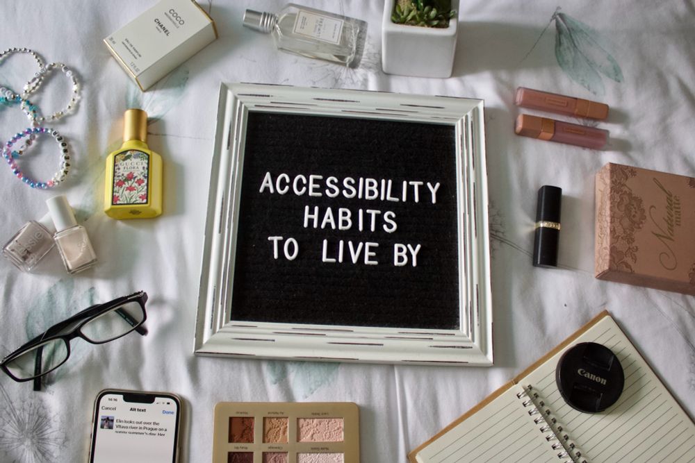 A black felt letterboard with a white rustic frame and white letters that spell out 'Accessibility habits to live by' is placed on a flat white surface with various items surrounding it, including a phone displaying Instagram's ALT text page, a notebook, a pair of black glasses, a few perfumes, a fake cactus and makeup products.