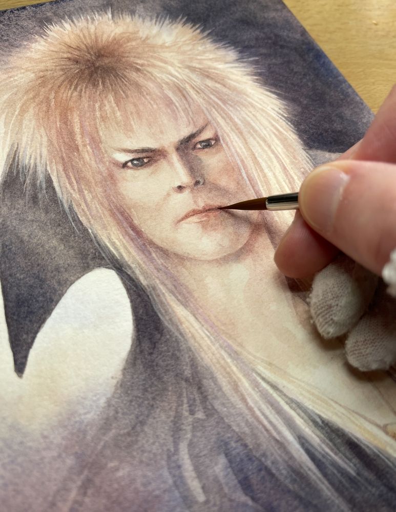 watercolour painting of jareth from labyrinth. You can see my hand and the paintbrush painting. He's got his signature blonde feathered mullet and a black high collar. 