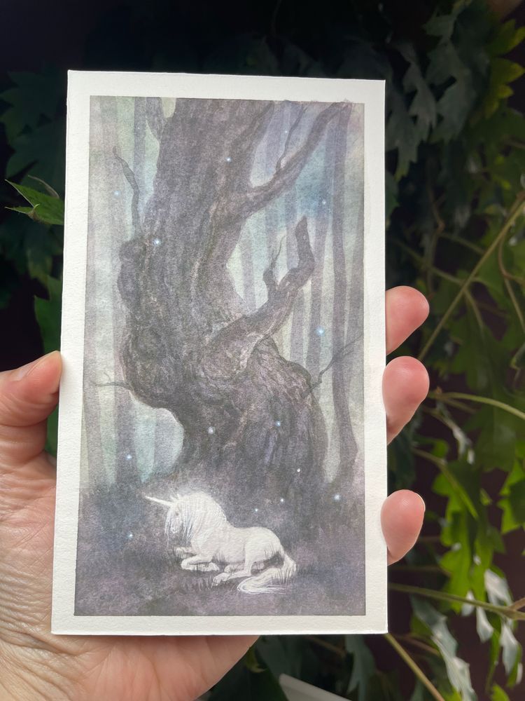Moody scene of a large twisted tree and a glowing unicorn resting beneath it. Small glowing lights float in the air around it 