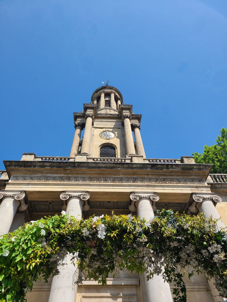 Photograph of a building with many columns and a clock tower seen from below. A fully blue sky is visible, along with a trellis covered in white flowers and leaves. 