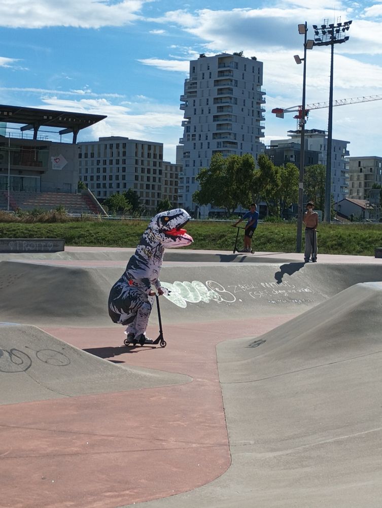 Photo d'un gars en train de faire de la trottinette sur le skatepark dans un costume de T-Rex gonflable 