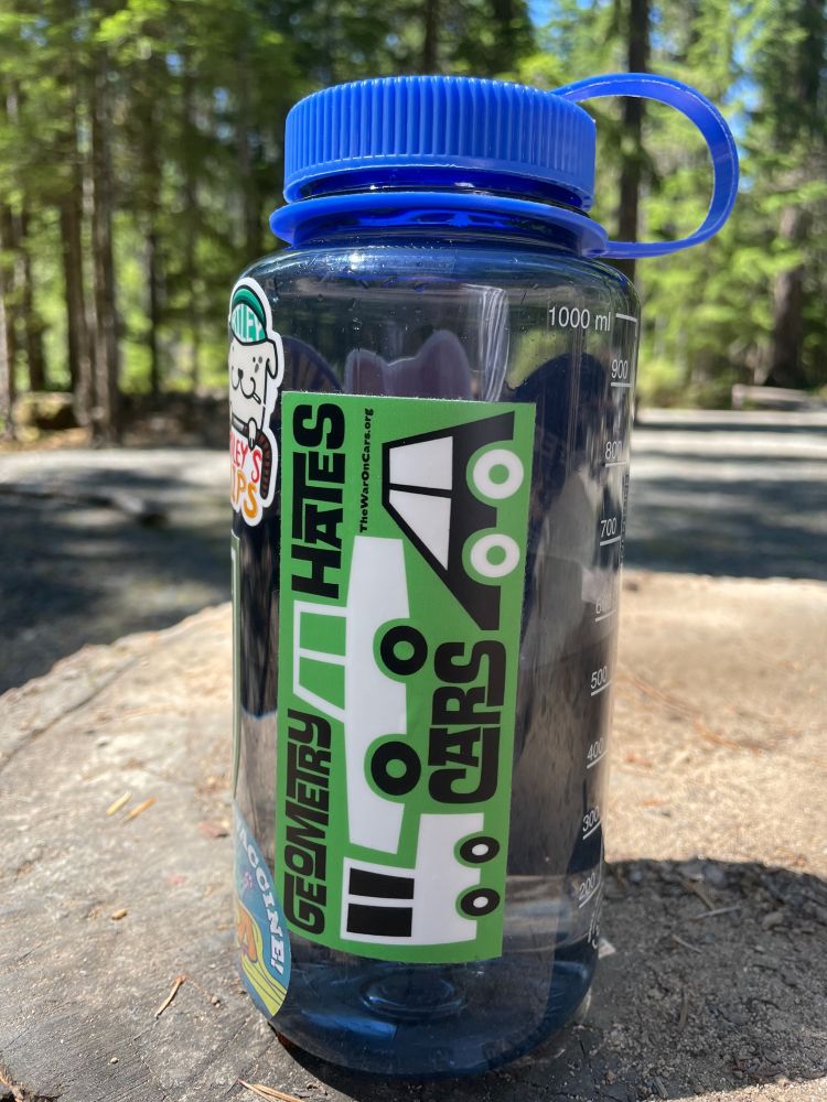 a photo of my fresh new nalgene bottle on a stump at a campsite. a green sticker is in the middle of it that says “geometry hates cars” with a graphic of some cars squished together (if only they fit together that well irl)