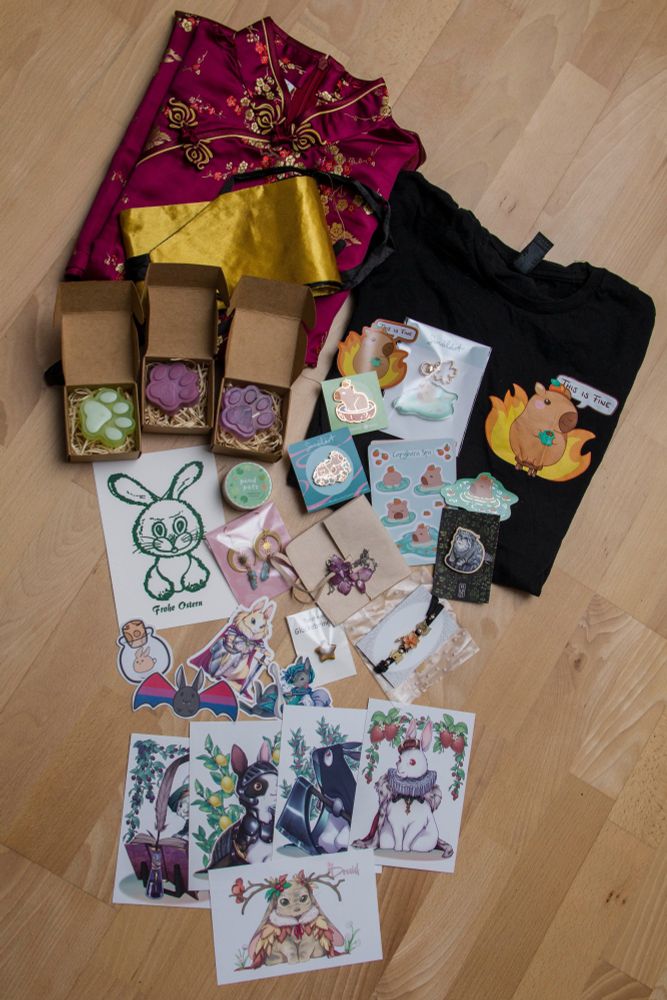 All different kinds of goodies:
* a Vietnamese dress
* three bars of paw-shaped soap
* a black shirt with a capybara version of the "This is fine"-meme
* capybara stickers, enamel pins and a capybara keychain
* a wooden pallas' cat pin
* a postcard with a bunny print saying "happy easter" (hand-printed at the stall of the German museum for printing in Leipzig)
* animal washi tapes
* earrings and two necklaces
* several cute stickers and postcards (including a bat with the bi-flag as wings and several bunnies inspired by DND / tabletop RPG characters)