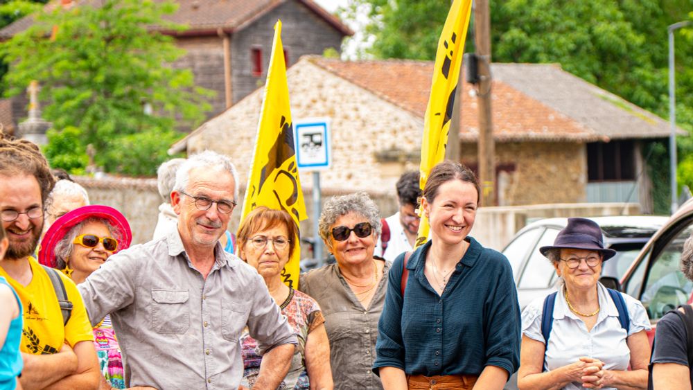 Plusieurs personnes souriantes rassemblées dans une rue de village. Deux drapeaux jaunes de la Confédération paysanne sont visibles. Au premier plan, Manon Meunier, députée de la Haute-Vienne et Philippe Babaudou ,co-Porte-Parole de la Conf 87, entourés de manifestant·es.