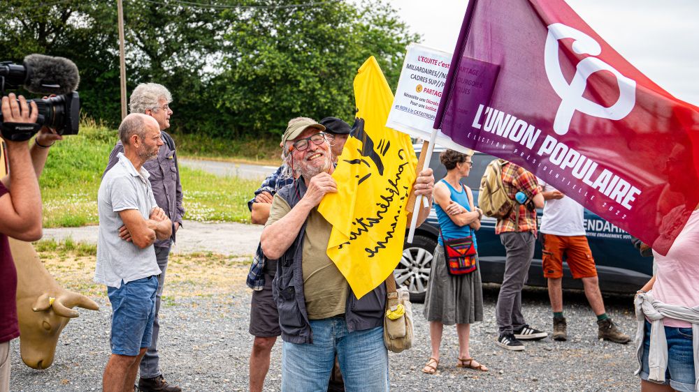 Un petit groupe de manifestant·es réuni·es en milieu rural. Un militant au centre sourit en tenant un drapeau jaune de la Confédération paysanne ainsi qu'un drapeau de L’Union Populaire. D'autres personnes discutent ou observent, tandis qu’un caméraman filme la scène.