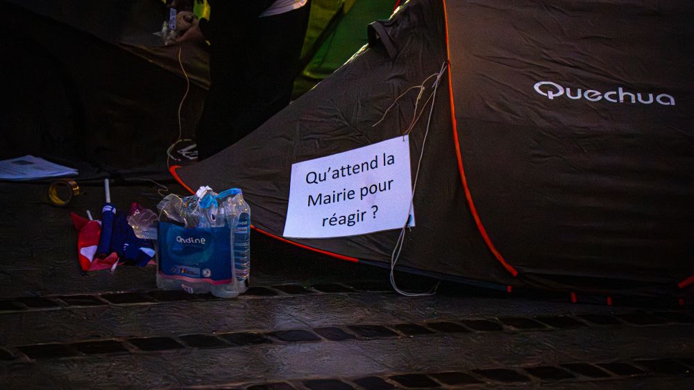 Une tente de camping installée devant la mairie de Limoges, avec une pancarte accrochée dessus où l’on peut lire : « Qu’attend la mairie pour réagir ? ». À côté, des bouteilles d’eau et des drapeaux pliés.