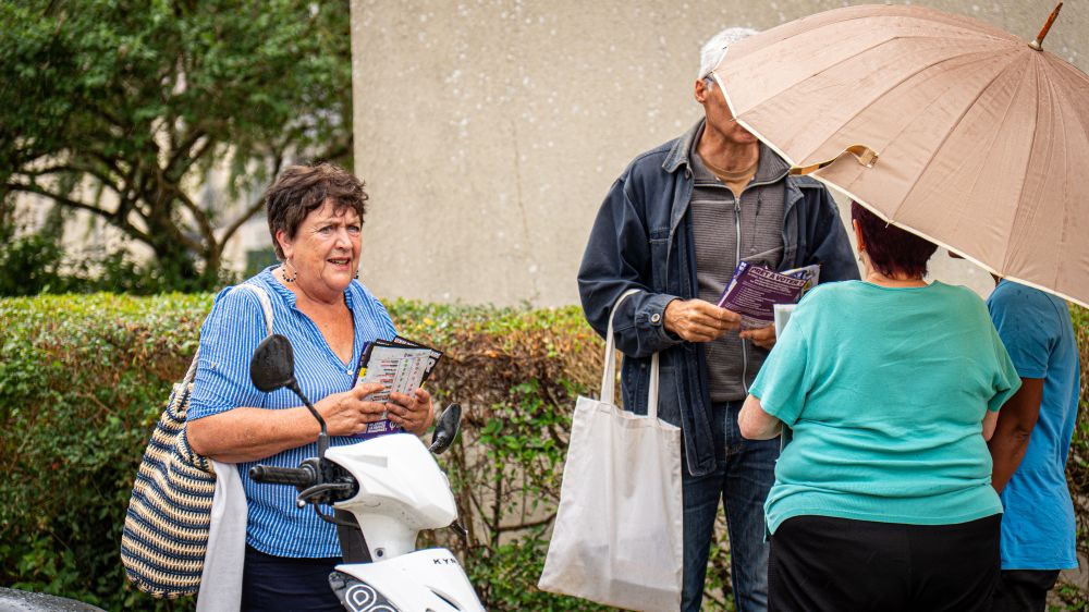 Scène de discussion sous la pluie : deux militant·es échangent avec des habitant·es abrité·es sous un grand parapluie beige. Une autre militante, debout près d’un scooter, distribue des tracts.