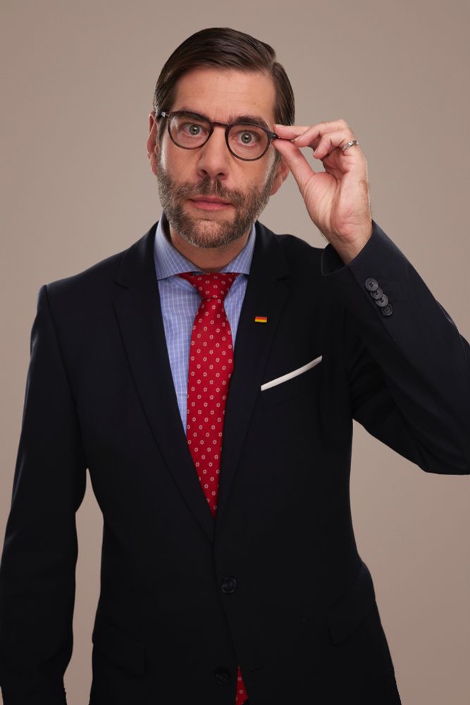 Claus von Wagner in einem feinen dunklen Anzug mit Einstecktuch und Deutschland Fahne am Revier, er stellt einen CDUler da - mit Brille.