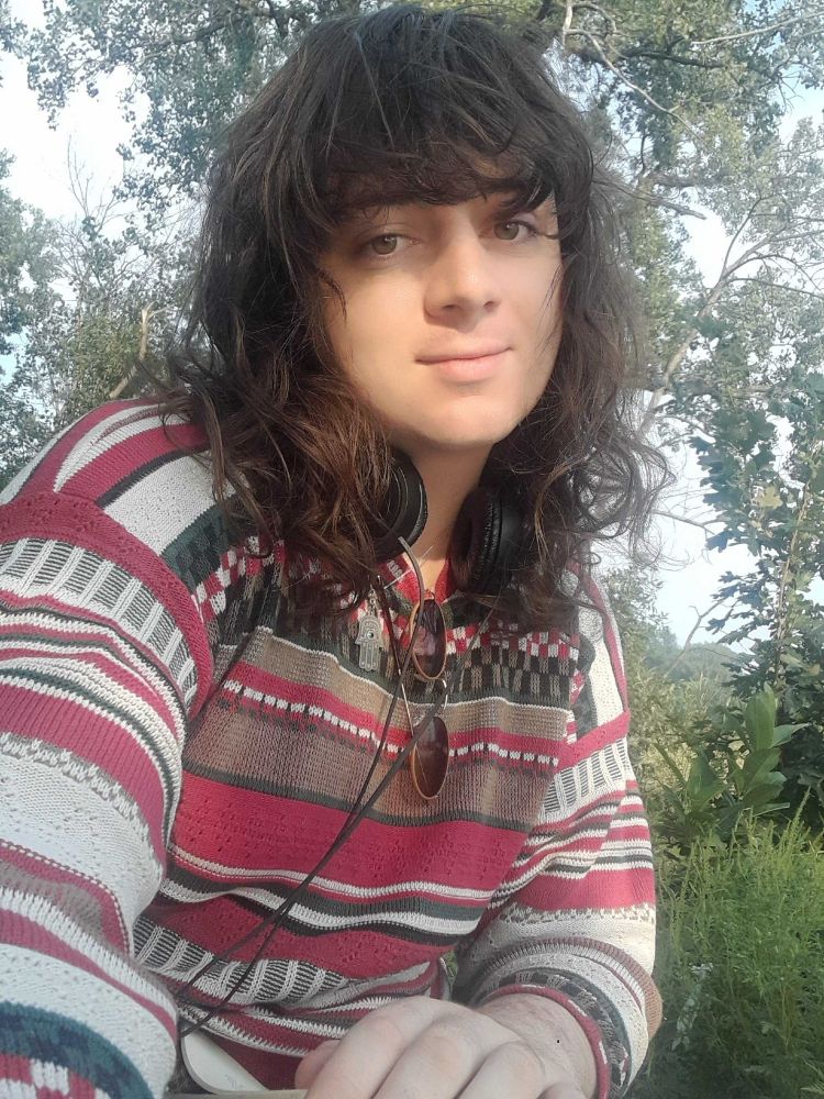 white lady with shoulder length hair and dark features in a colorful knit sweater with headphones around her neck a hamsa pendant and sunglasses on her neckline sat in a wood and looking at the viewer placidly smiling. i think