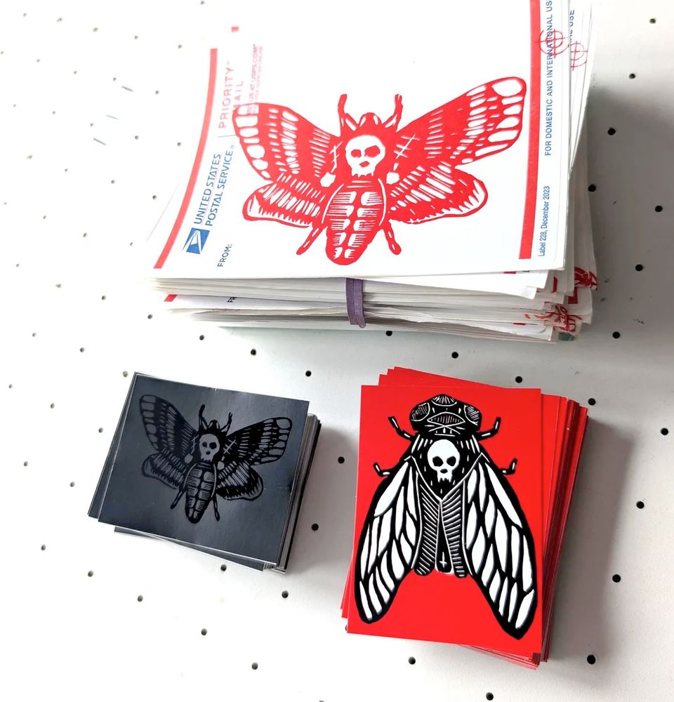 The contents of a typical sticker pack: 3 different stickers by west coast graffiti & wheatpaste artist DEBUG - hand screen printed label 228s with red moth, black-on-black limited edition moth vinyl stickers, red eggshell security stickers. 

Photographed on my printmaking vacuum table from above