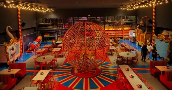 Um restaurante pizzaria temático de circo com mesas brancas e cadeiras vermelhas, o chão colorido em vermelho, azul e amarelo e tem uma grande bola enfeitada no meio