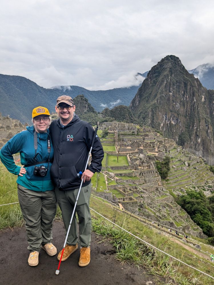 Bryanna and her husband stand by Machu Picchu ruins in Peru. He has a white cane. 