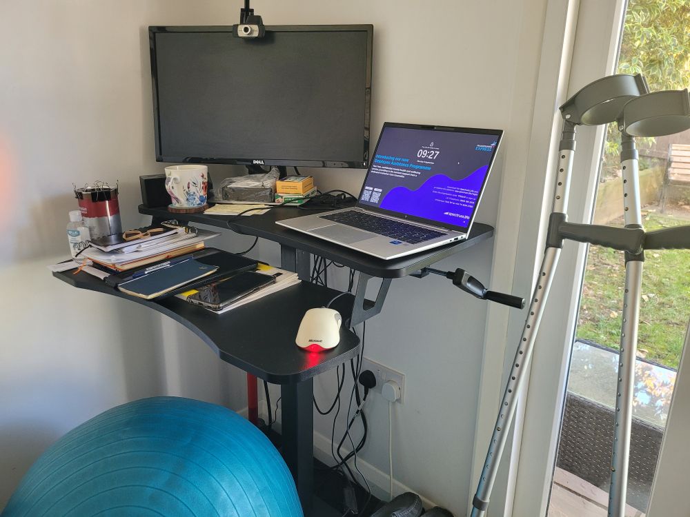 A standing desk in the lowered (seated) position. A turquoise Swiss Ball underneath and a pair of crutches lean against the window frame to the left of the desk.