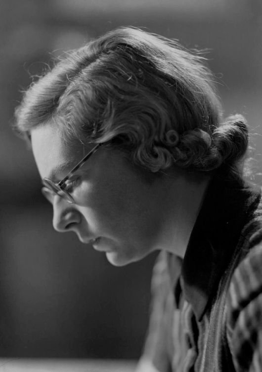 Black and white photograph of composer Elizabeth Maconchy. It is taken from the left hand side - she is looking down, wearing glasses, and has short hair curled at the bottom. She wears a dark striped shirt.