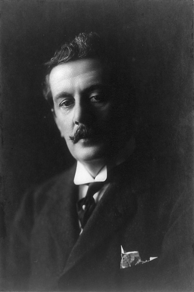 Black and white photograph of Puccini. He has a thick moustache and dark hair, and while facing the camera his face is half in shadow. He wears a dark suit.