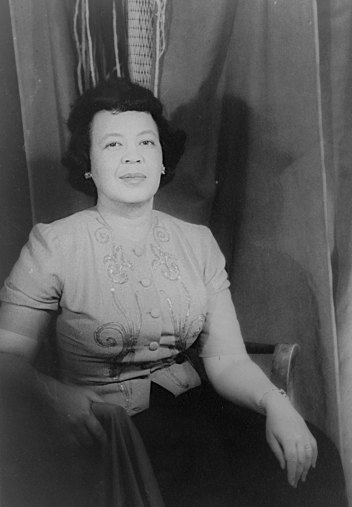 Black and white photograph of compoer Margaret Bonds. She is wearing a pale sequinned top, and a dark skirt. She sits in front of a curtain.