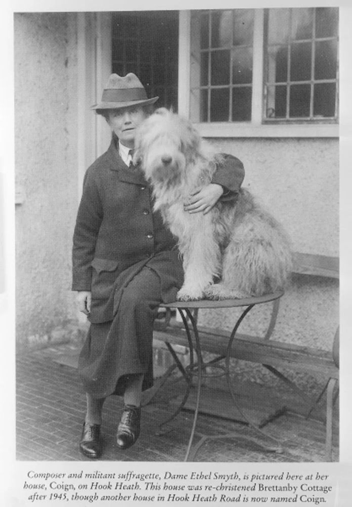 Black and white photograph of the composer Ethel Smyth with her sheepdog Pan. She is wearing a hat and long coat with a shirt, tie, and long skirt with flat patent shoes. They are sitting on her outdoor table, by the window of her house Coign on Hook Heath.