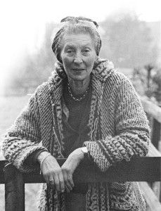 Black and white photograph of composer Elizabeth Poston. She is standing in a foggy field leaning on a fence, wearing a long cardigan and hair tied back with a scarf.