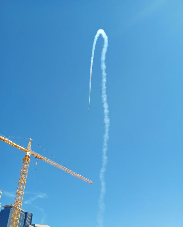 A blue-sky day in Seattle.

A yellow industrial crane.
A sliver of the Emerald City skyline.

A Blue Angel jet. 
Boeing F/A-18 Super Hornet.

An arching, white contrail.

A beautiful summer day on the Elliott Bay waterfront.