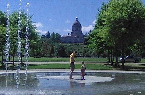 A dad and two children stand in the dry zone of the water fountain in downtown Olympia.

Water is in the air to the left.

The state Capitol Building is in the background.

There are ample green trees and grass 