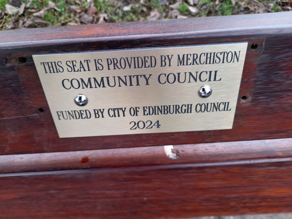 A metal plaque affixed to a refurbished wooden bench. The plaque reads "This seat is provided by merchiston community council. Funded by City of Edinburgh Council. 2024"