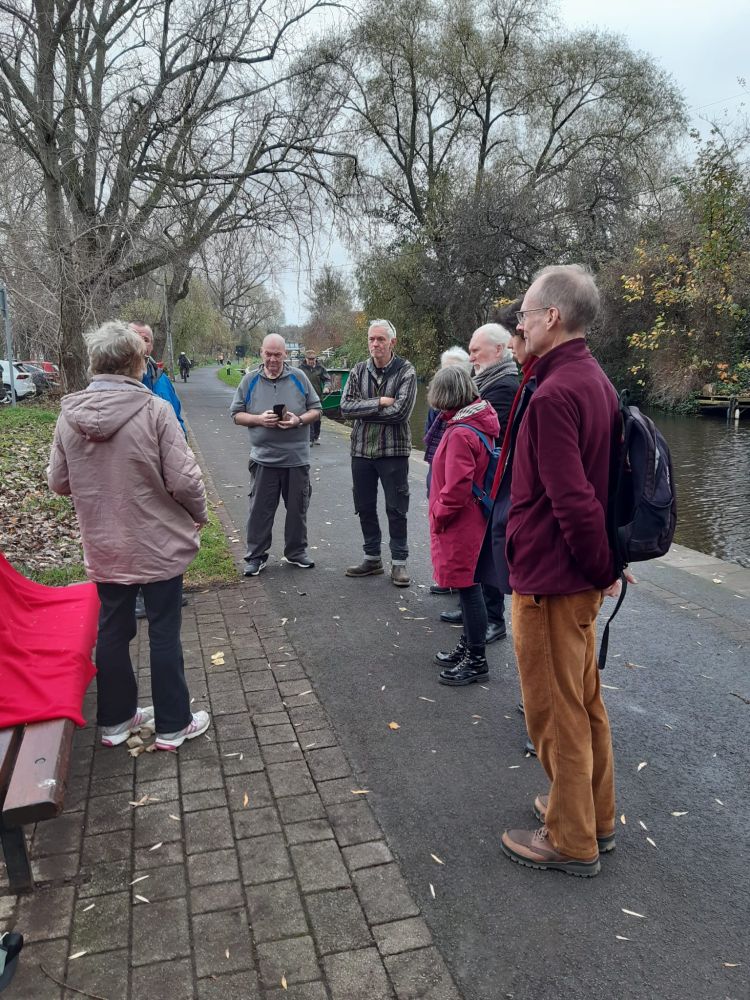 A group of people stand on a canal towpath as a newly restored bench is unveiled