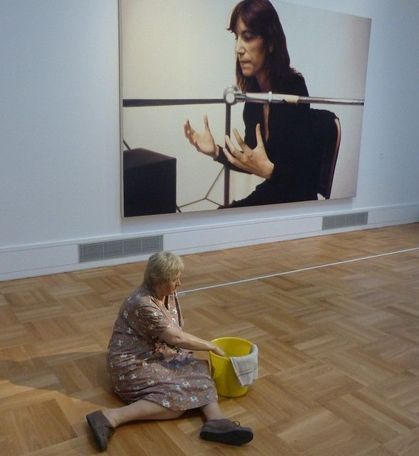 Das Bild zeigt Skulptur von Duane Hanson sitzt, die eine Putzfrau darstellt. (Staatsgalerie Stuttgart).
Die Skulptur ist Teil des hyperrealistischen Werks von Duane Hanson, einem amerikanischen Künstler, der für seine lebensechten Statuen von Alltagsmenschen bekannt ist. 
Hanson verwendete Polyesterharze mit Glasfaser, um Abgüsse von echten Menschen herzustellen, und bemalte die Figuren dann mit großer Detailgenauigkeit. 
Dieses Werk, das eine Putzfrau darstellt, ist ein Beispiel für Hansons Fähigkeit, gewöhnliche Szenen und Charaktere des amerikanischen Lebens einzufangen. 
Die Skulptur befindet sich in einem Ausstellungsraum und interagiert auf subtile Weise mit der realen Person im Vordergrund, was die Grenzen zwischen Kunst und Realität verwischt.