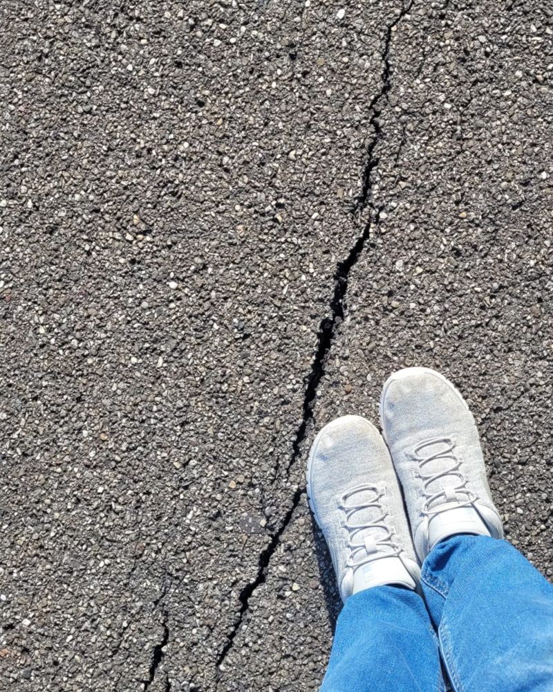 Dieses Bild zeigt eine Ich-Perspektive, bei der die Beine und Füße einer Person im unteren Teil des Bildes sichtbar sind. Die Person trägt hellblaue Jeans und hellgraue oder weiße Sportschuhe mit weißen Schnürsenkeln. Die Füße stehen auf einem dunklen Asphaltboden, der eine deutlich sichtbare, unregelmäßige Risslinie aufweist, die sich von der unteren Bildmitte diagonal nach oben und leicht nach links zieht. Der Asphalt scheint aus vielen kleinen grauen und schwarzen Körnchen zu bestehen, was eine raue Textur erzeugt. Das Licht fällt von oben auf die Szene, was zu einem leichten Schattenwurf unter den Füßen führt und die Textur des Asphalts hervorhebt.
