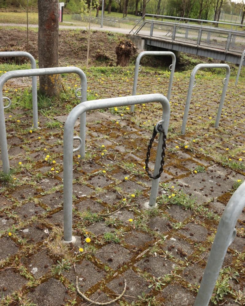 Das Bild zeigt einen Außenbereich, der als Fahrradabstellplatz dient. Es sind mehrere leere, U-förmige Fahrradständer aus Metall zu sehen, die in regelmäßigen Abständen auf einem gepflasterten Boden angeordnet sind.
Im Vordergrund und in der Mitte des Bildes ist ein einzelner Fahrradständer zu sehen, an dem eine dunkle, rostige Kette hängt, die an einem Ende mit einem Schloss oder einem ähnlichen Verschluss versehen ist. Die Kette ist um den oberen Teil des Bügels gewickelt. Das dazugehörige Fahrrad fehlt.  