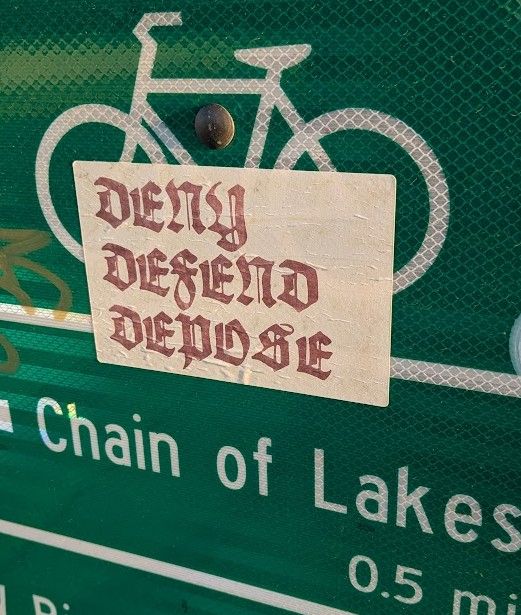 Sticker on a street sign reads, "Deny, Defend, Depose." 