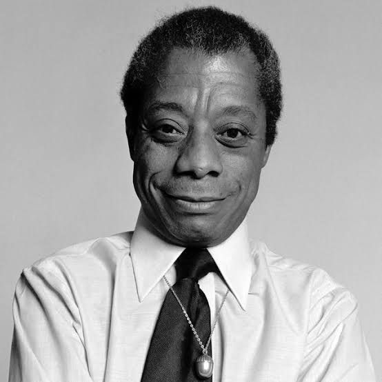 A photgraph of James Baldwin. He is wearing a white collared shirt and a dark tie and an amulet on a chain around his neck. He is smiling and looking direct at the camera. 