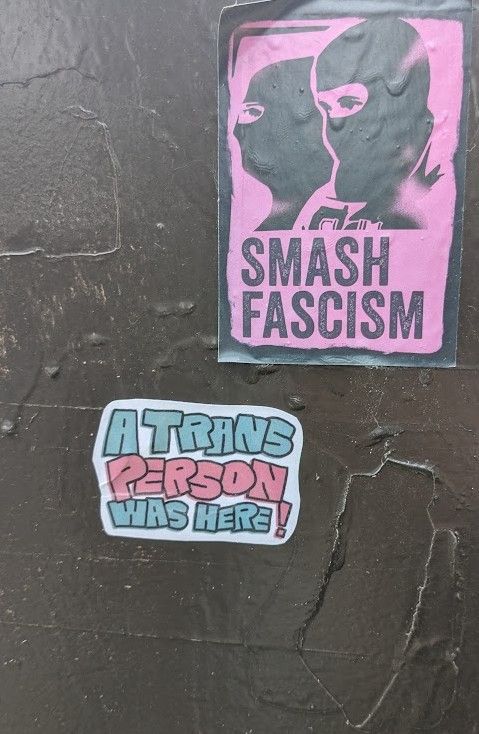 Two stickers on the side of a public electric utility box. One reads, "A trans person was here!" The other has an image of two people in ski masks and reads, "Smash Fascism." 